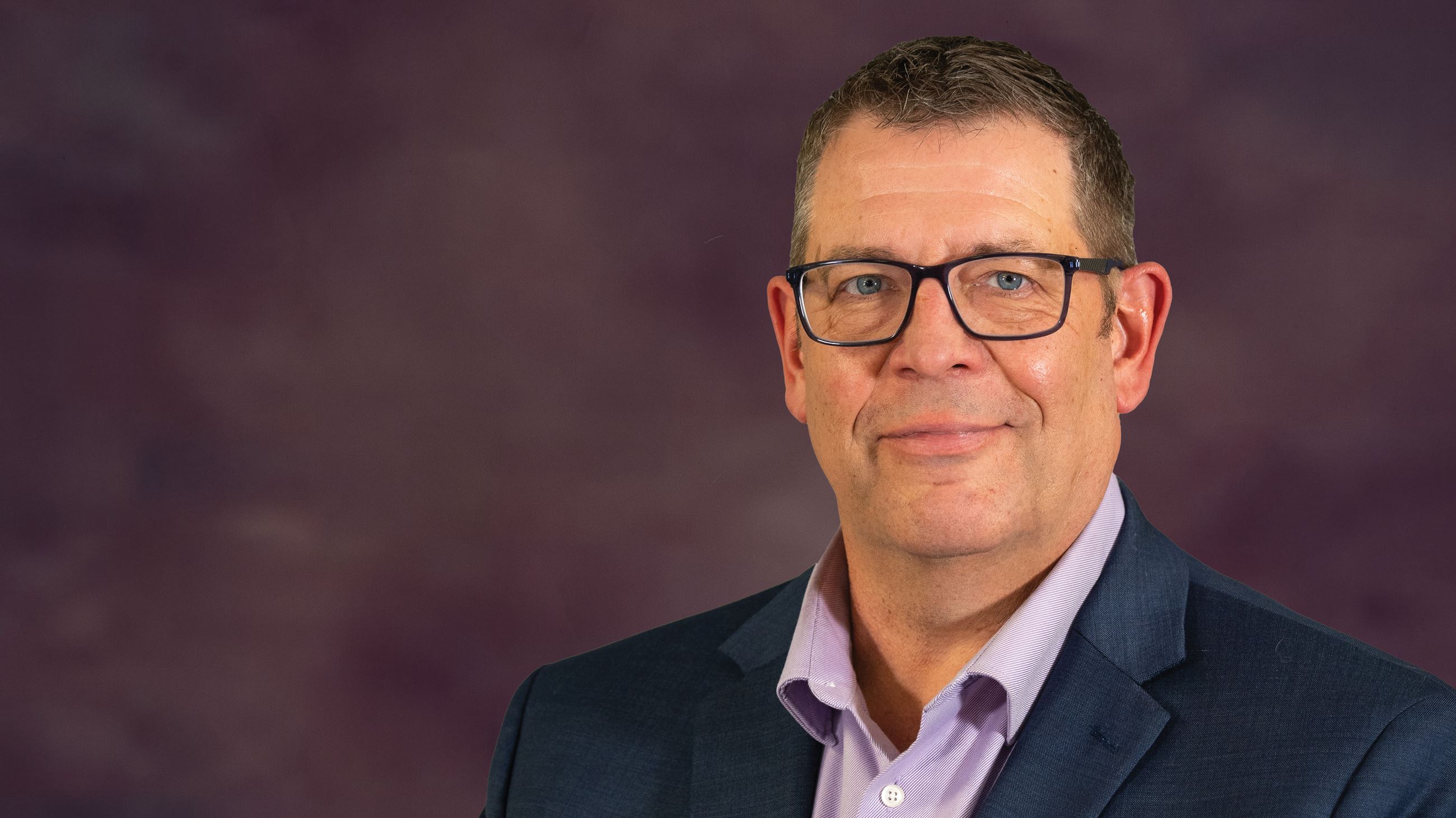 A man in glasses and a suit and tie poses for a photo in front of a purple background.