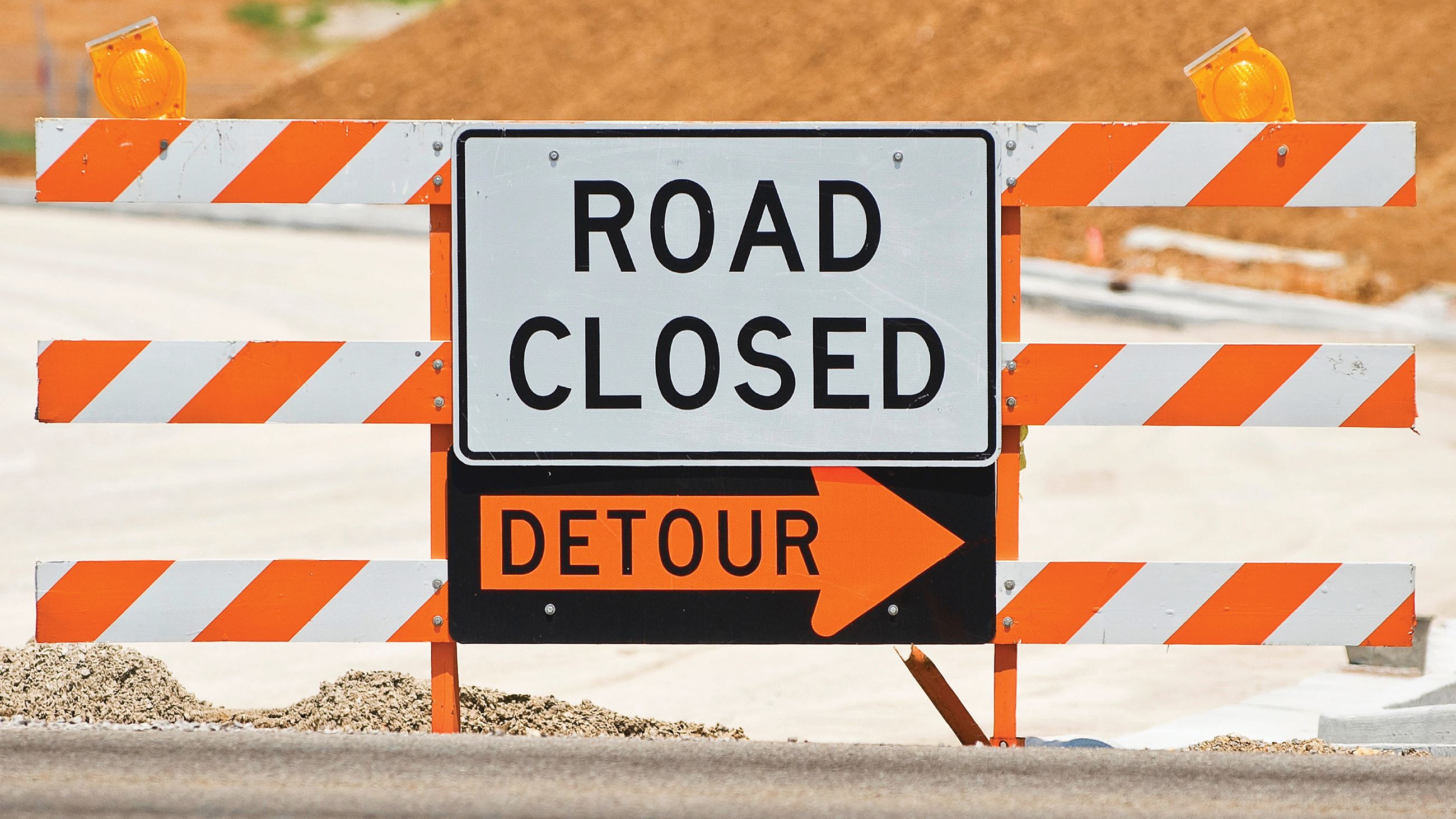 An orange and white striped barricade sits in front of a road with a detour sign marked on it.