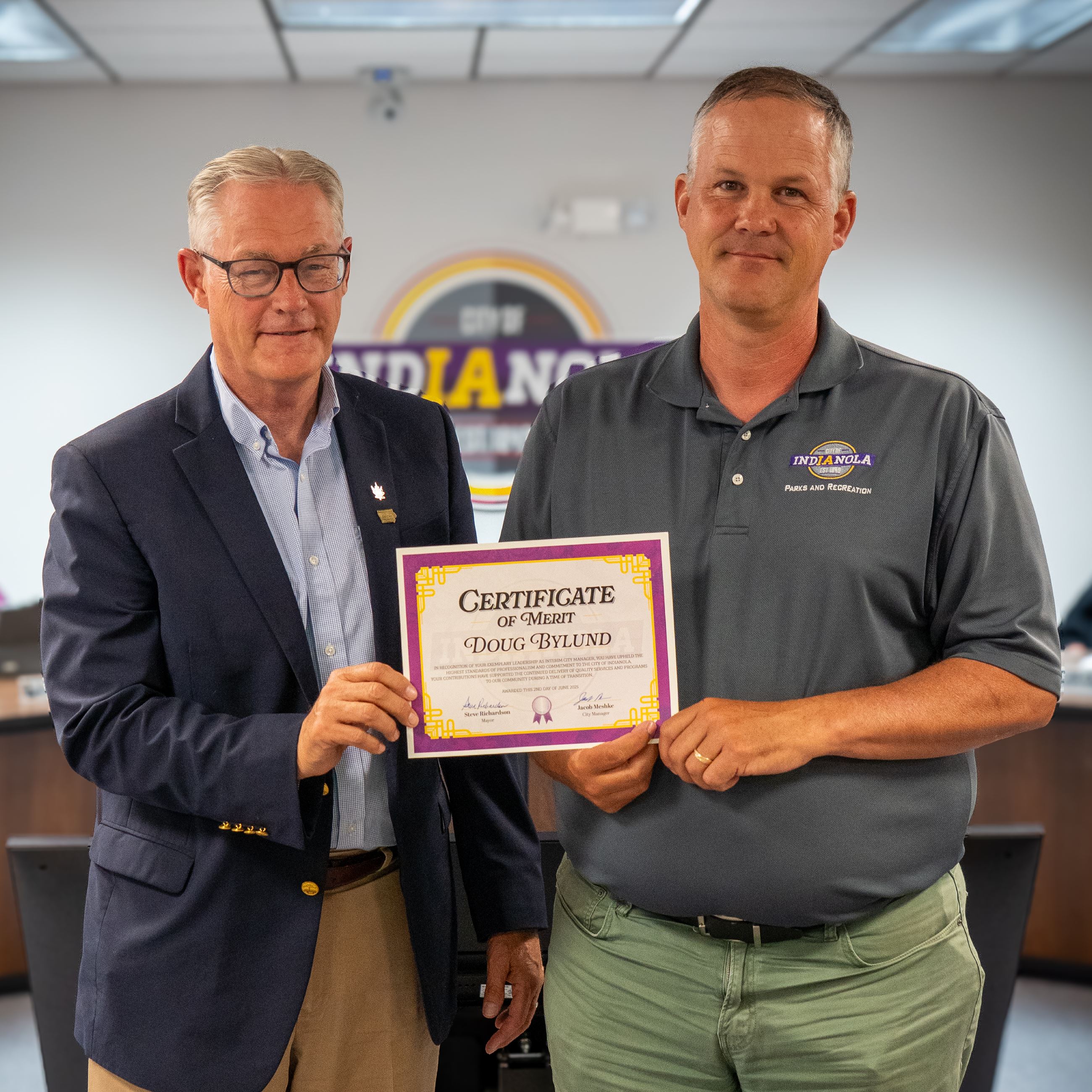 A man wearing a suit and glasses and another man wearing a gray polo hold an award certificate. 