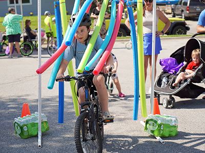 A boy wearing a bike helmet rides his bicycle through a colorful curtain of pool noodles